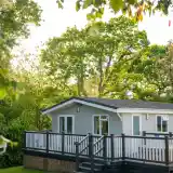 A modern gray cabin with a white deck surrounded by lush green trees and sunlight filtering through the leaves.