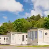 Two modern holiday homes are nestled among lush green trees under a bright blue sky. The homes feature large windows and light-colored exteriors, with a welcoming atmosphere for vacationers.