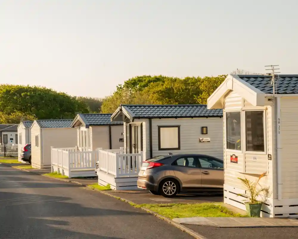 A row of small, white holiday homes with porches and sloped roofs lined along a paved road. Several cars are parked in front of the homes, with trees in the background under a clear sky.