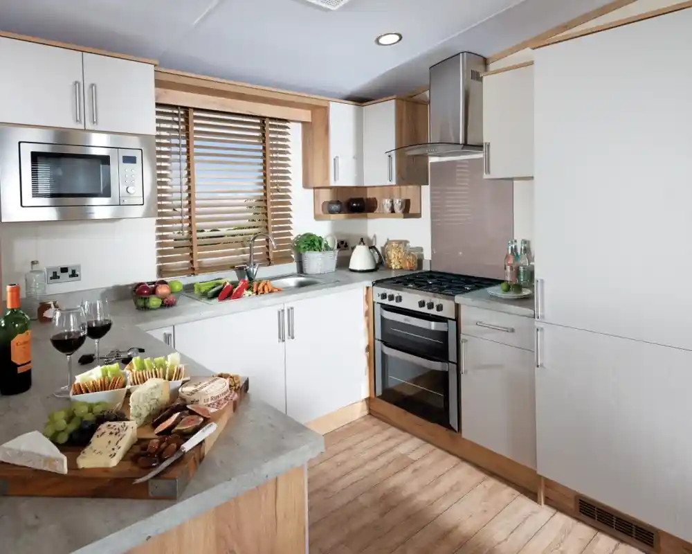 A modern kitchen with light wood accents and white cabinetry. The Countertop features a wooden serving platter with various cheeses, fruits, and wine glasses. A stove, microwave, and kettle are visible, along with a window covered by wooden blinds, allowing natural light to stream in.