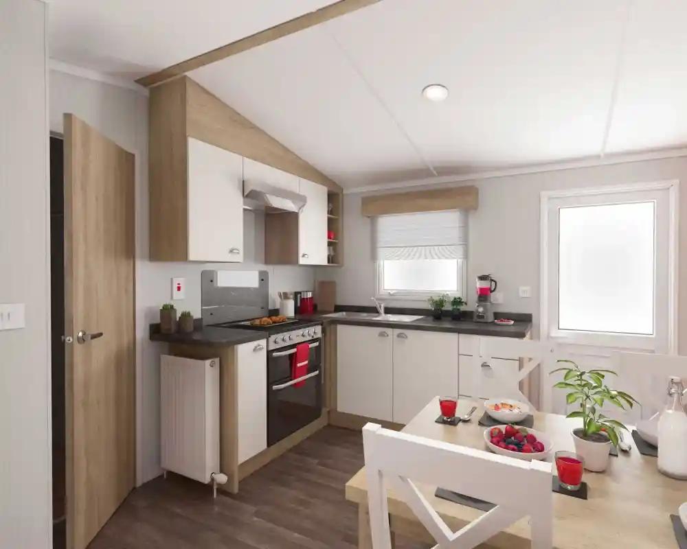 A modern kitchen featuring light wood cabinets, a black countertop, and stainless steel appliances. A dining area with a white table and chairs is visible, adorned with red glasses and a bowl of mixed fruit. Natural light streams in through a window.