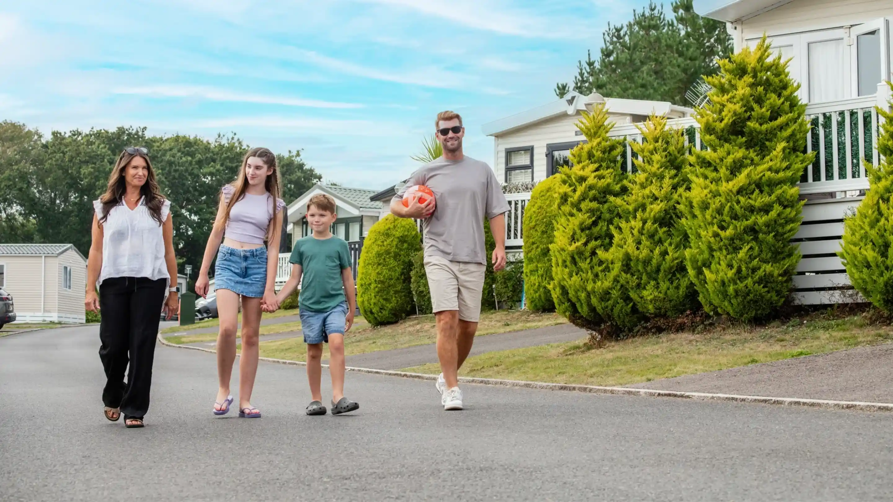 A family strolls down a street lined with neatly trimmed hedges and homes. A man holds a soccer ball, accompanied by a woman and two children, all smiling and enjoying their time together. The sky is clear with a hint of clouds.