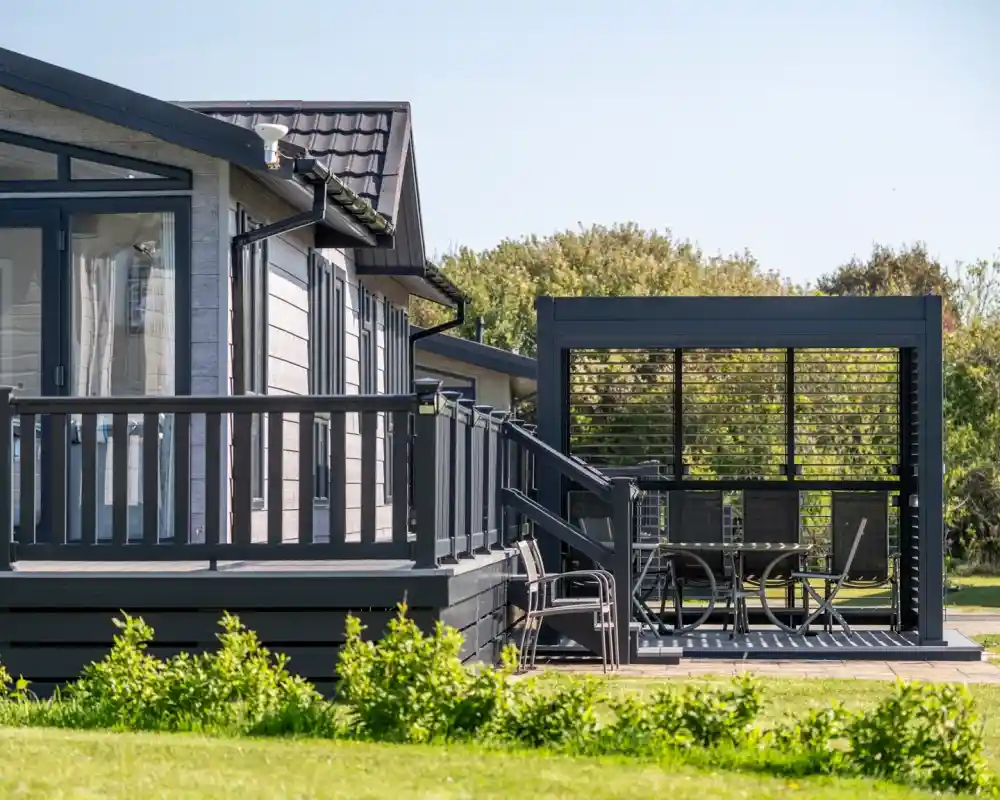 A modern wooden cabin with a deck and a shaded outdoor seating area is surrounded by lush greenery. The cabin features large windows and a dark exterior, while the patio has a dining set.