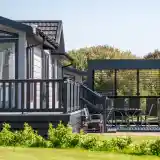 A modern wooden cabin with a deck and a shaded outdoor seating area is surrounded by lush greenery. The cabin features large windows and a dark exterior, while the patio has a dining set.