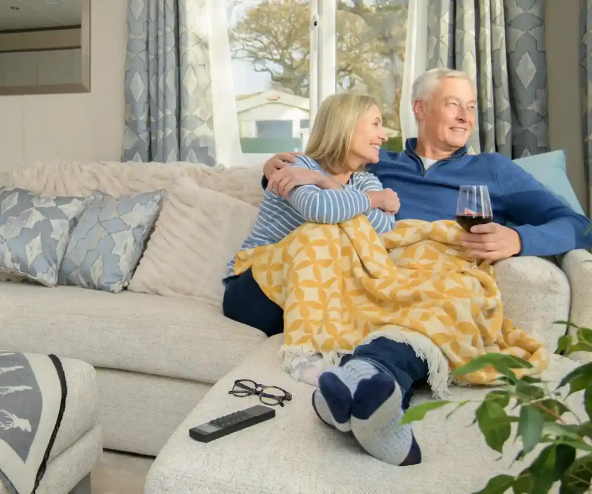 A couple stands on a balcony with a modern home in the background. The man and woman are smiling, dressed casually, with green grass and a landscaped area surrounding the house. The sky is clear and blue.