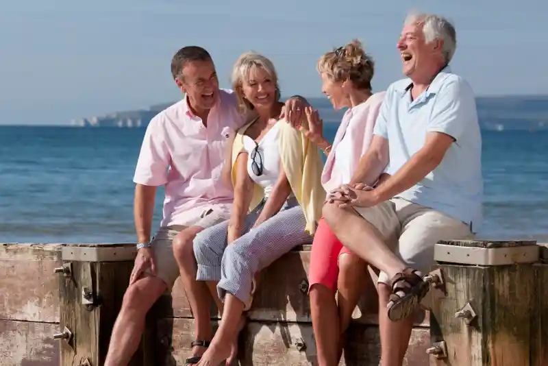 Four people sit on a wooden pier by the beach, laughing and enjoying each other's company. They are dressed in casual summer clothing, with the ocean and a clear sky in the background.