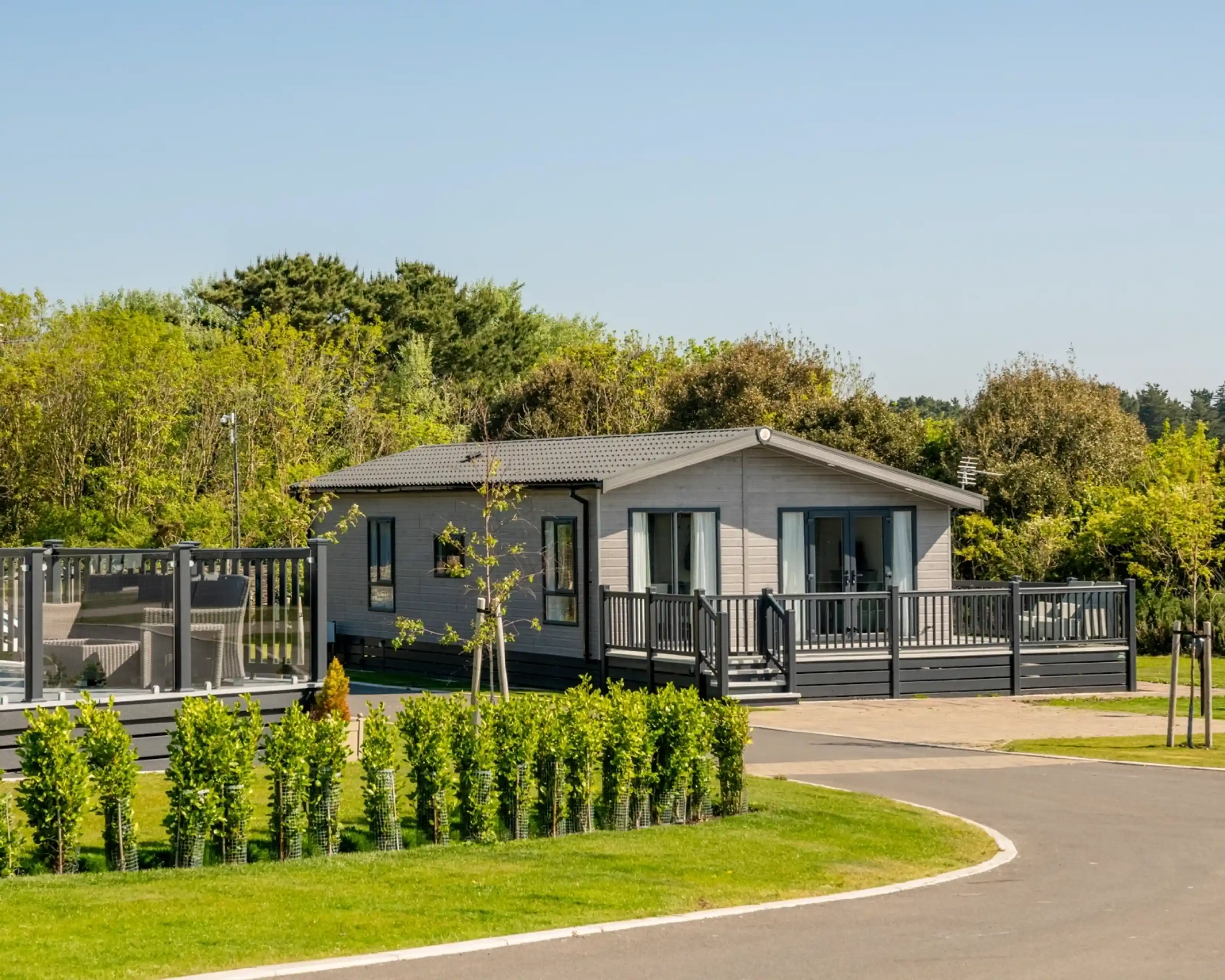 A modern gray house with a deck, surrounded by greenery and neatly trimmed hedges, located within a park-like setting. A paved path curves around the property under a clear blue sky.