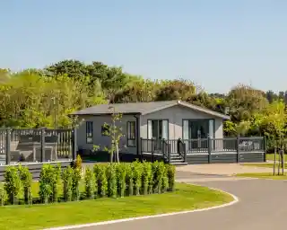 A modern gray house with a deck, surrounded by greenery and neatly trimmed hedges, located within a park-like setting. A paved path curves around the property under a clear blue sky.