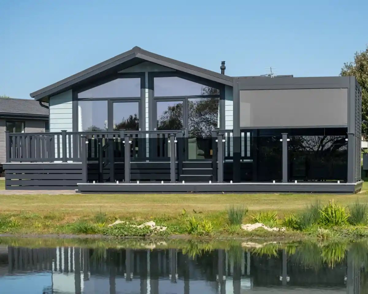 A modern, gray house with large windows and a deck, situated by a small pond surrounded by greenery. Clear blue sky is overhead.