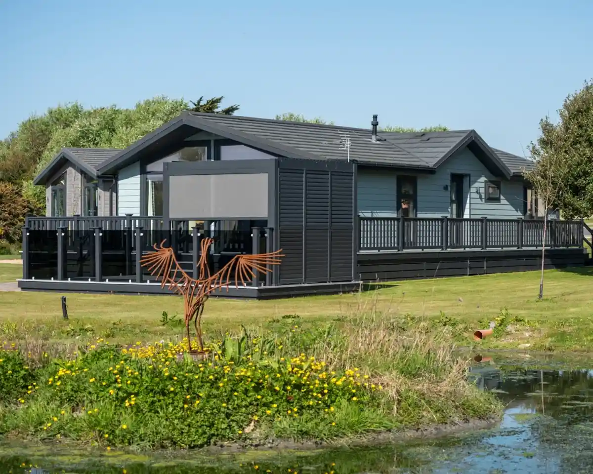 A modern, wooden cabin with a wraparound deck set in a green landscape. There is a small pond with yellow flowers in the foreground and a decorative metal bird sculpture in the yard.