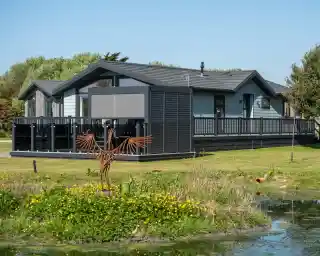 A modern, wooden cabin with a wraparound deck set in a green landscape. There is a small pond with yellow flowers in the foreground and a decorative metal bird sculpture in the yard.