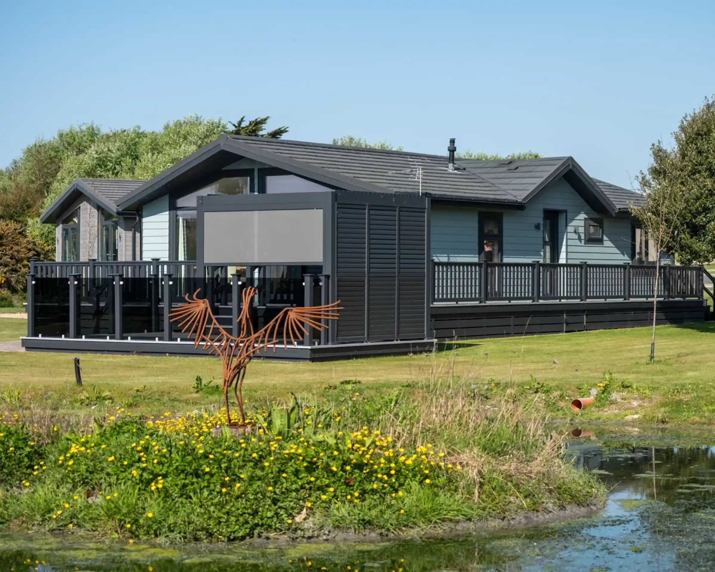 A modern, wooden cabin with a wraparound deck set in a green landscape. There is a small pond with yellow flowers in the foreground and a decorative metal bird sculpture in the yard.