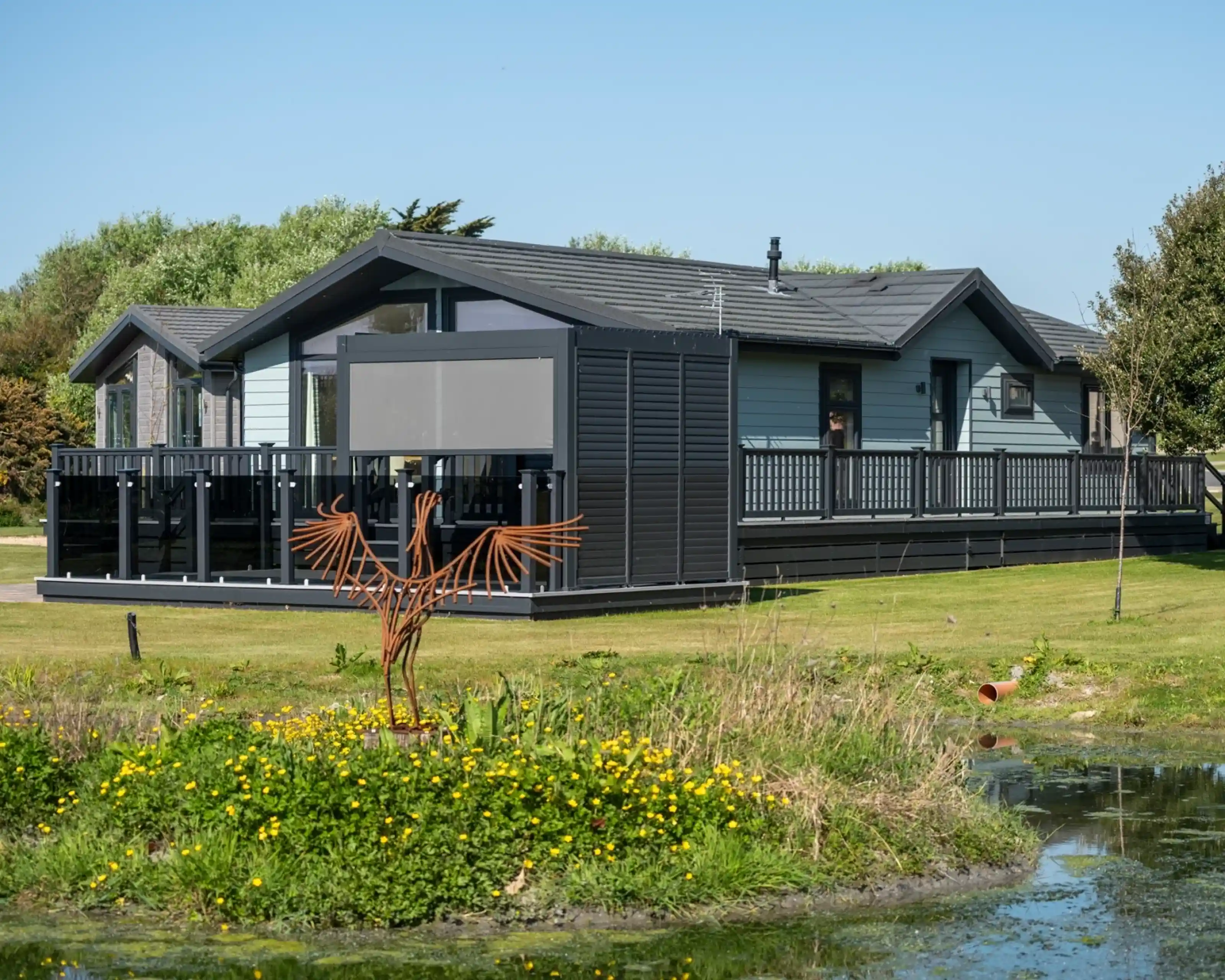 A modern, wooden cabin with a wraparound deck set in a green landscape. There is a small pond with yellow flowers in the foreground and a decorative metal bird sculpture in the yard.