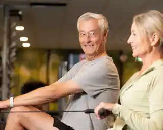 A older man and woman are smiling while using rowing machines in a gym. The setting is bright, with gym equipment visible in the background.