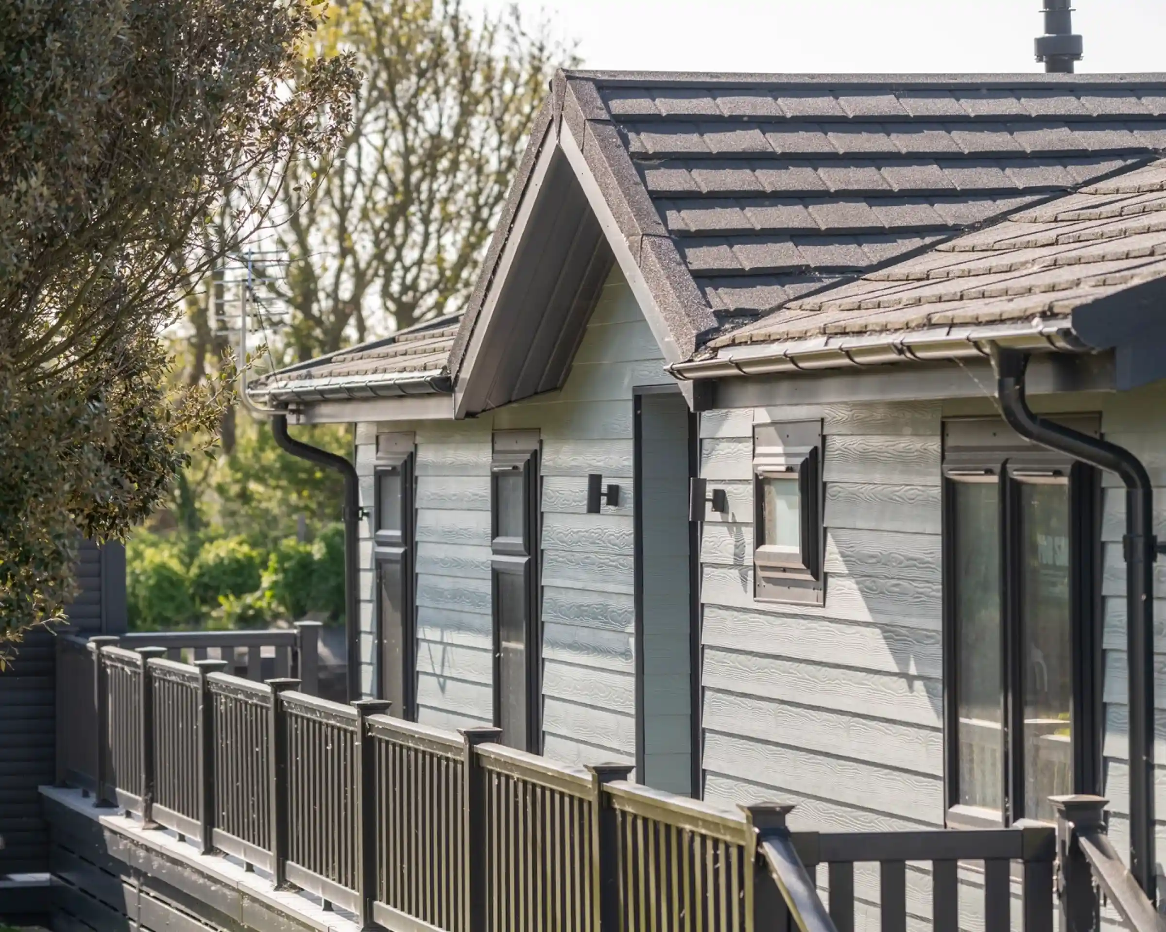 A modern cabin with a sloped roof and wooden balcony, surrounded by greenery. The exterior features light gray wood paneling and stylish wall-mounted lights.
