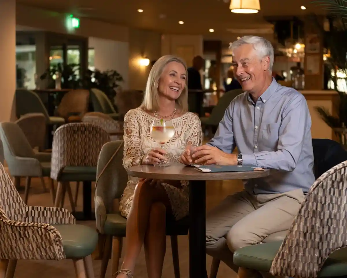 A smiling couple sits at a round table in a cozy lounge, enjoying drinks. The woman wears a stylish cream dress and the man is in a light blue shirt. They appear engaged in a lively conversation, surrounded by warm lighting and modern decor.
