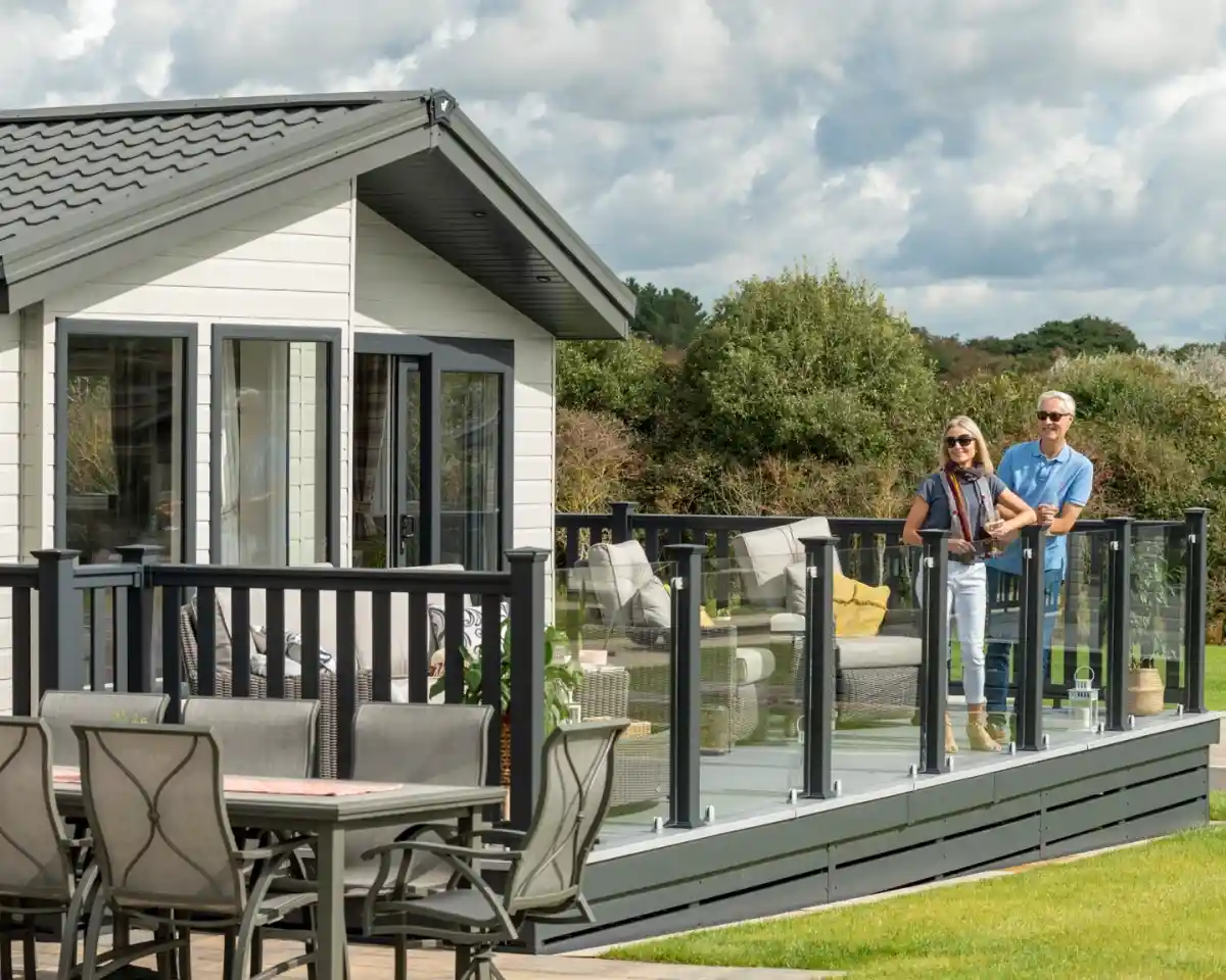 A couple stands on a stylish deck of a modern cabin, overlooking a grassy area. The woman wears sunglasses and a stylish outfit, while the man is casually dressed. Outdoor furniture is visible in the background, surrounded by greenery under a partly cloudy sky.
