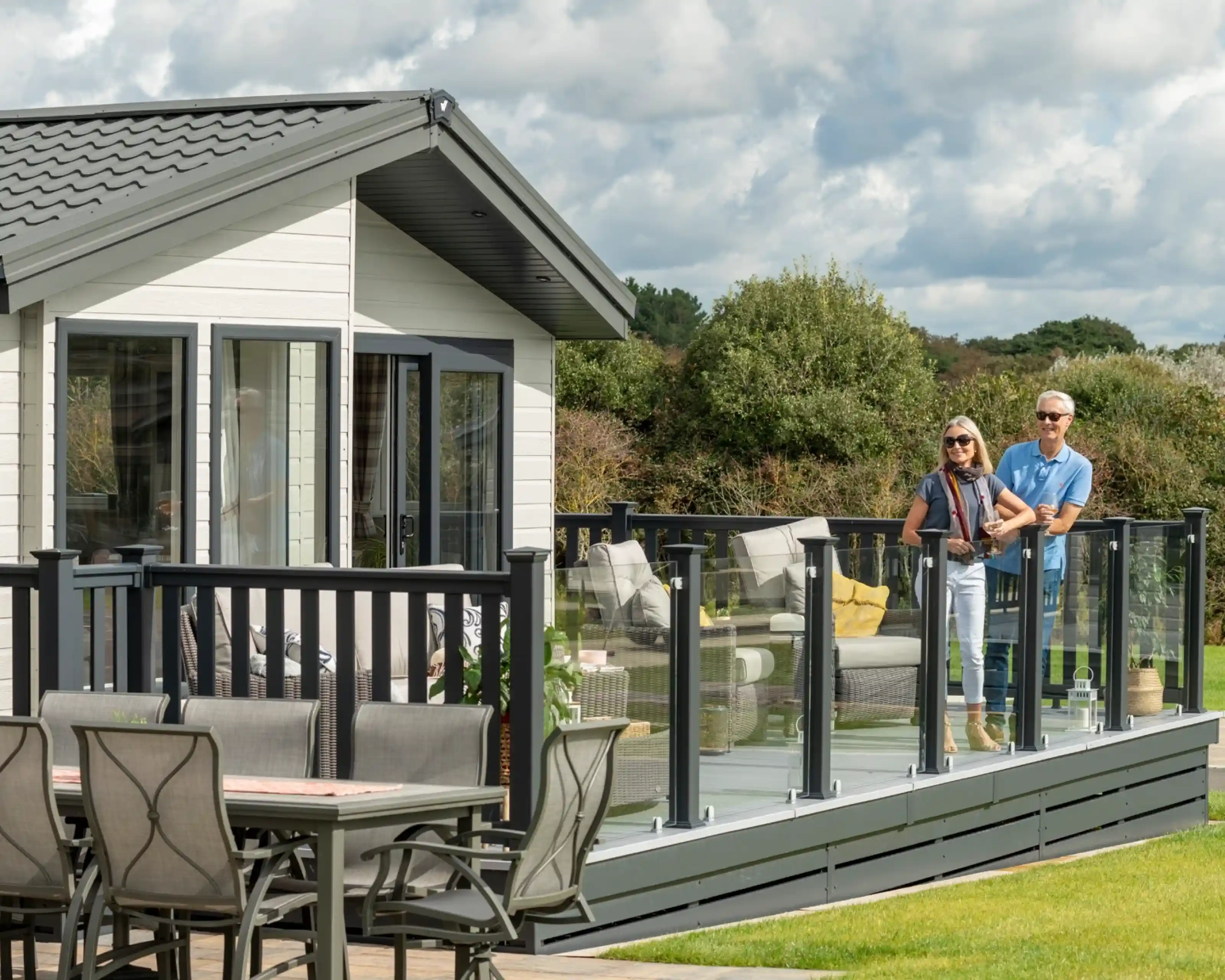A couple stands on a stylish deck of a modern cabin, overlooking a grassy area. The woman wears sunglasses and a stylish outfit, while the man is casually dressed. Outdoor furniture is visible in the background, surrounded by greenery under a partly cloudy sky.