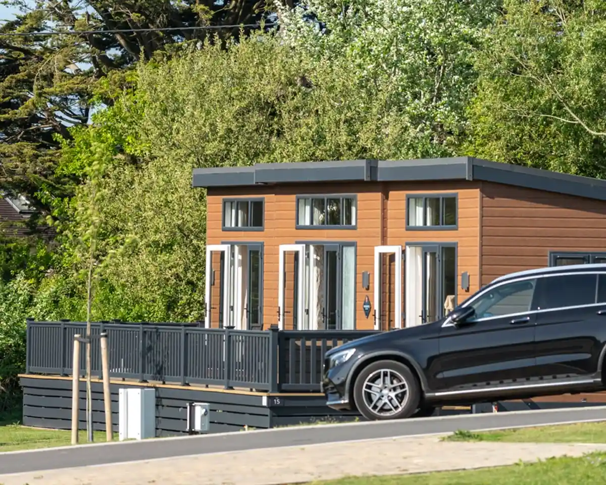 A modern cabin with large front windows and a wooden deck is surrounded by greenery. A black SUV is parked in front on a paved driveway.
