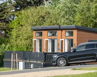A modern cabin with large front windows and a wooden deck is surrounded by greenery. A black SUV is parked in front on a paved driveway.