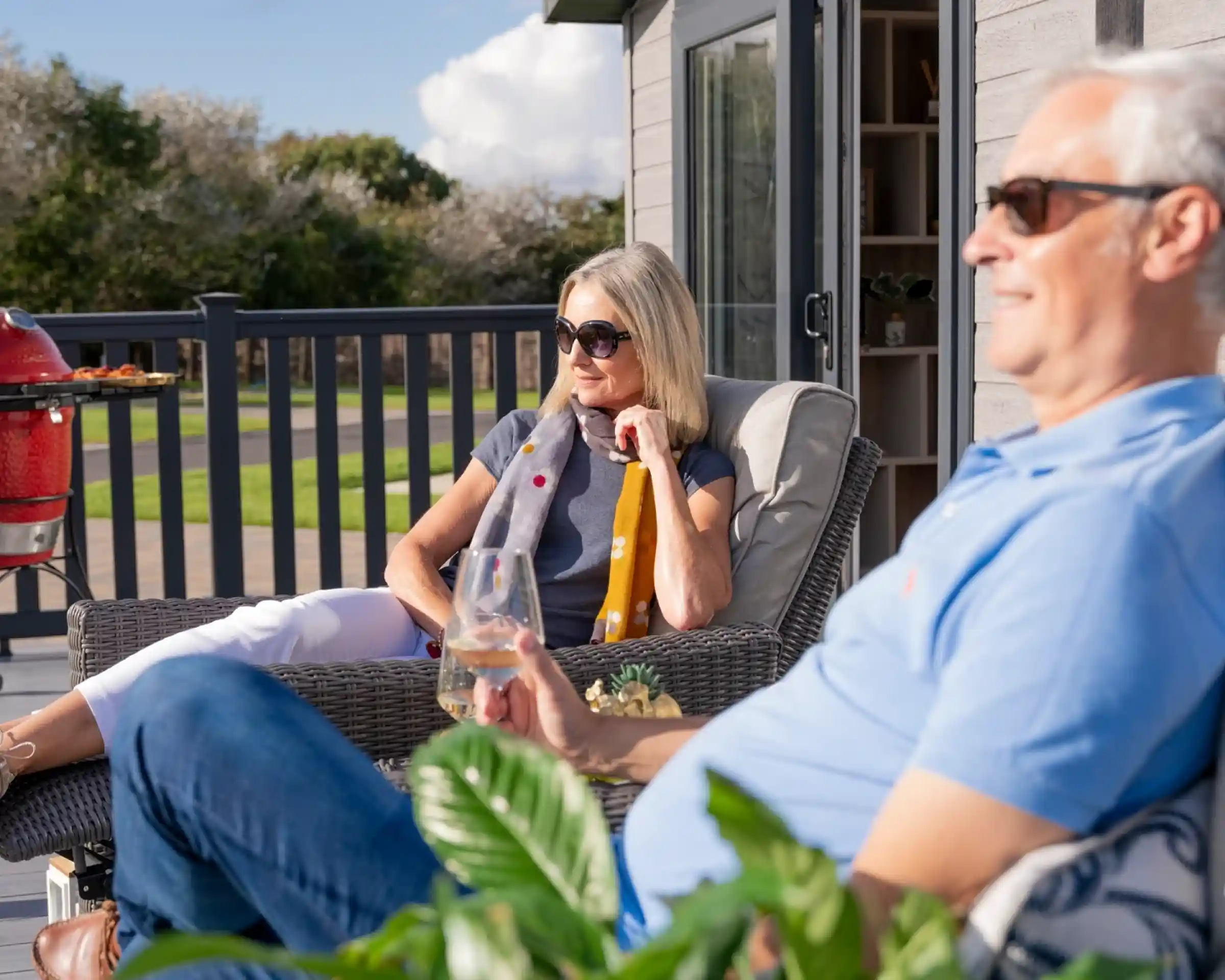A relaxed couple enjoys a sunny day on a patio. The woman, wearing sunglasses and a grey top, sits thoughtfully with a drink, while the man, in a blue polo shirt, reclines nearby, also in sunglasses. Lush greenery surrounds them, and a grill is visible in the background.