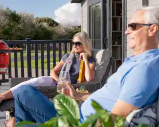 A relaxed couple enjoys a sunny day on a patio. The woman, wearing sunglasses and a grey top, sits thoughtfully with a drink, while the man, in a blue polo shirt, reclines nearby, also in sunglasses. Lush greenery surrounds them, and a grill is visible in the background.
