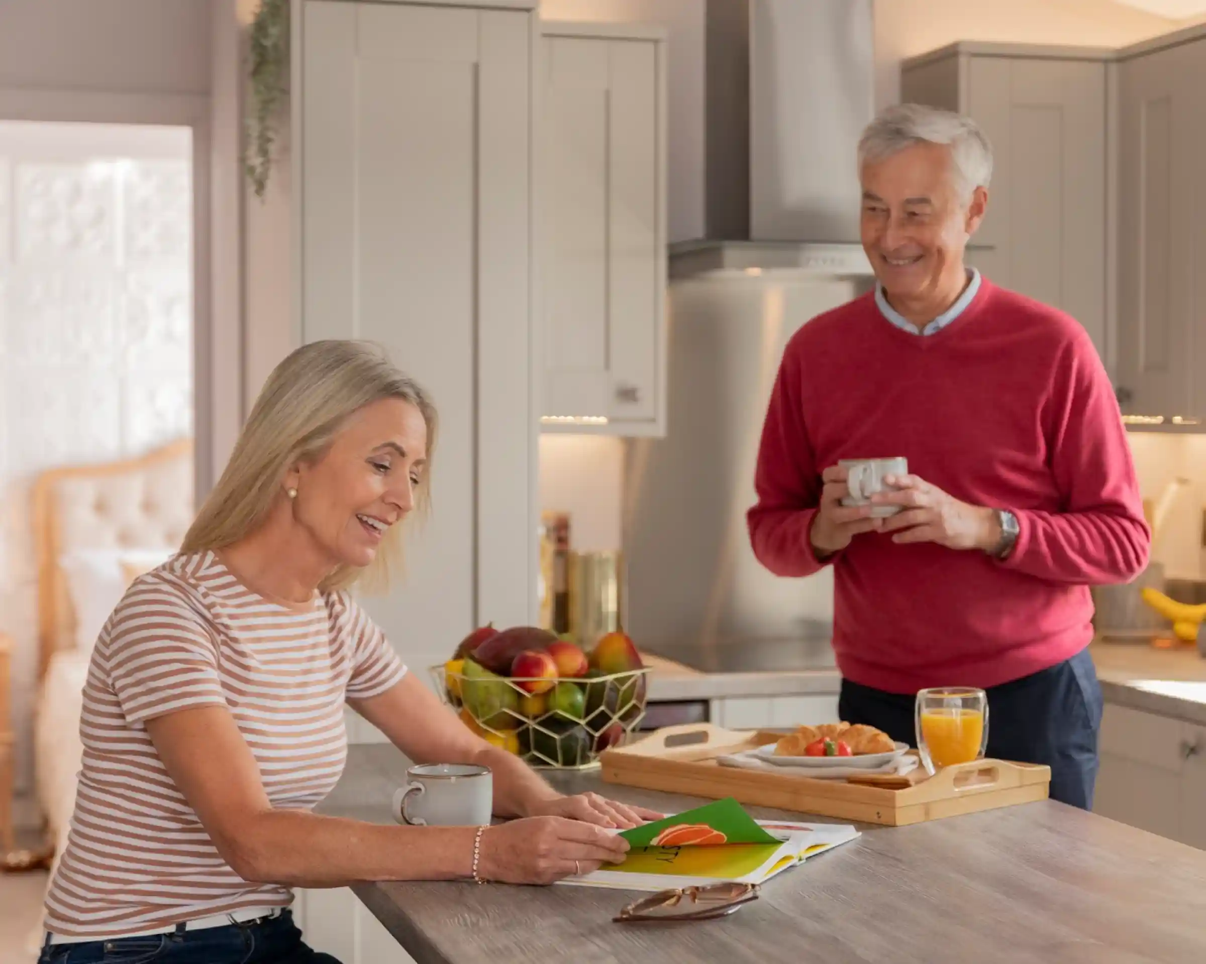 A woman with long blonde hair smiles while looking at a colorful booklet on a kitchen island. A man with gray hair and wearing a red sweater stands nearby, holding a cup and smiling at her. A fruit bowl and a tray with food can be seen in the background.