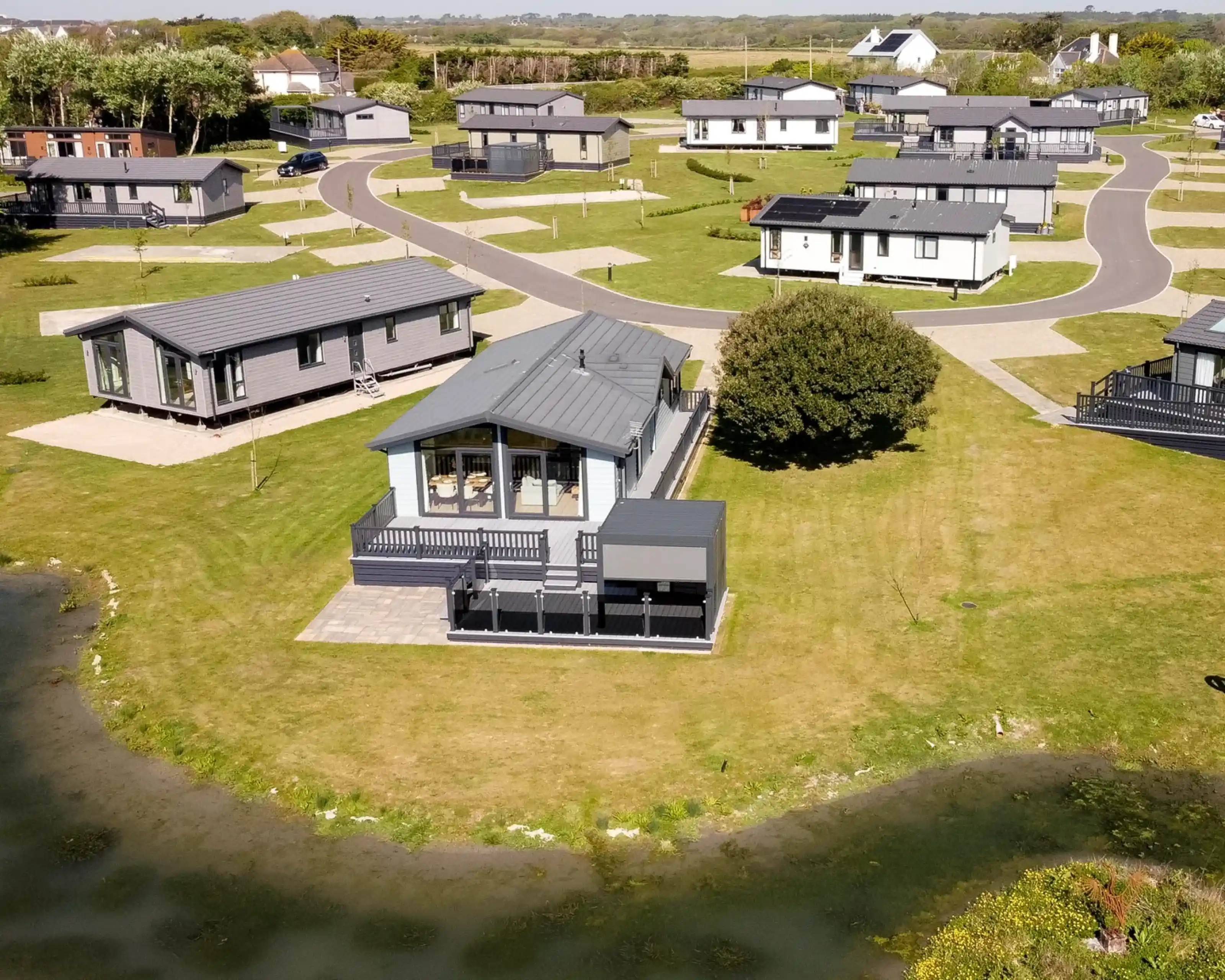 Aerial view of a holiday park featuring several modern lodges arranged along curved roads, surrounded by green grass and a small body of water.