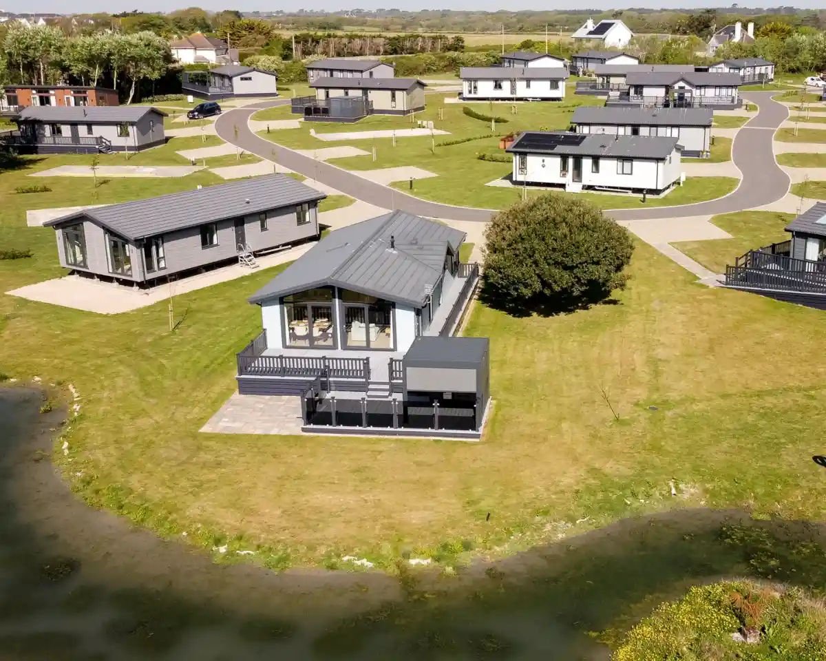 Aerial view of a holiday park featuring several modern lodges arranged along curved roads, surrounded by green grass and a small body of water.