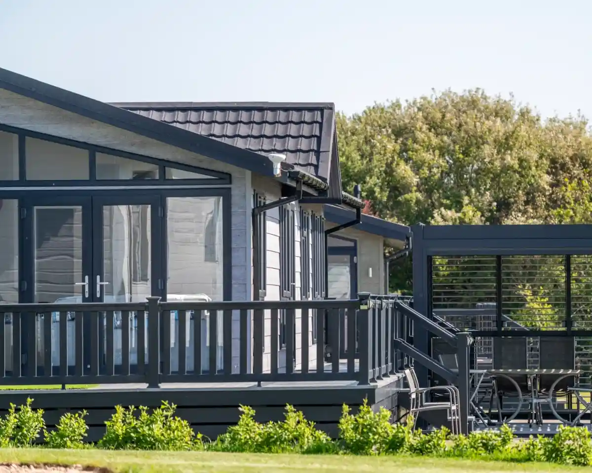 A modern dark gray house with large windows and a porch, surrounded by greenery. The design features a sloped roof and a deck with seating.