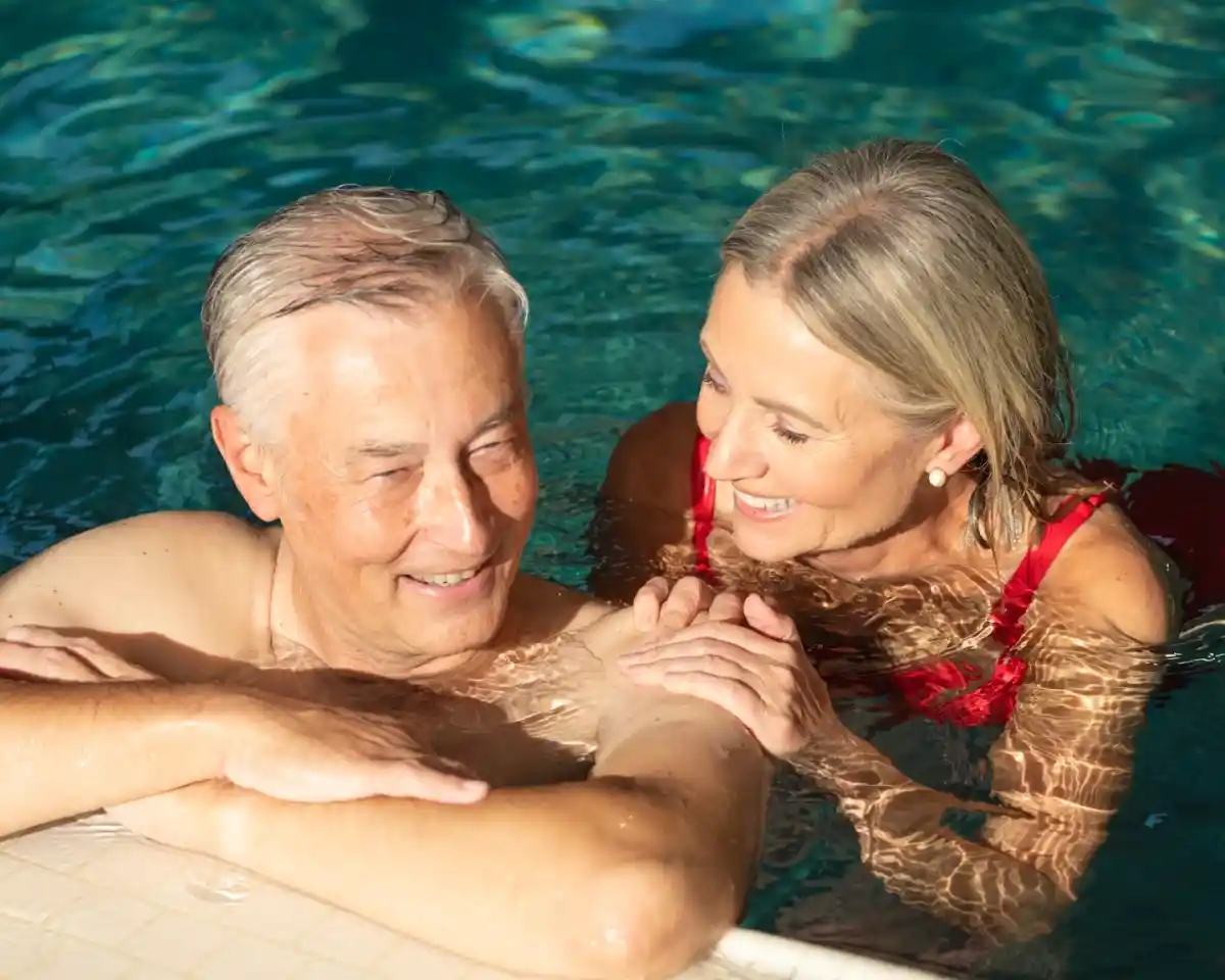 An older couple smiles and interacts playfully in a swimming pool, with sunlight glistening on the water's surface. The man is shirtless, while the woman wears a red swimsuit and has her hand gently placed on his arm.
