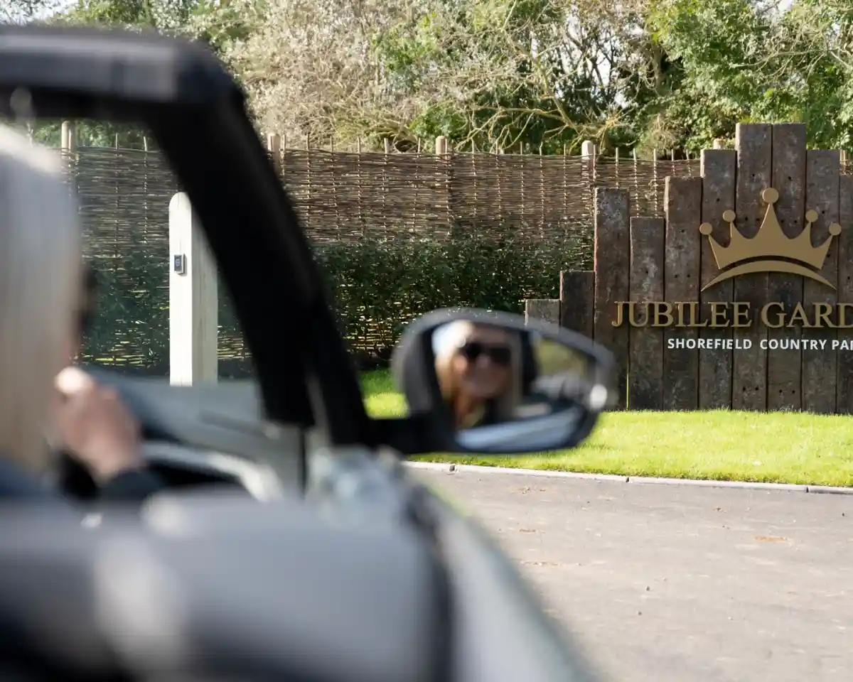 A person is seated in a convertible car, looking towards a sign that reads "Jubilee Gardens, Shorefield Country Park." The sign is surrounded by a wooden fence and green landscaping.
