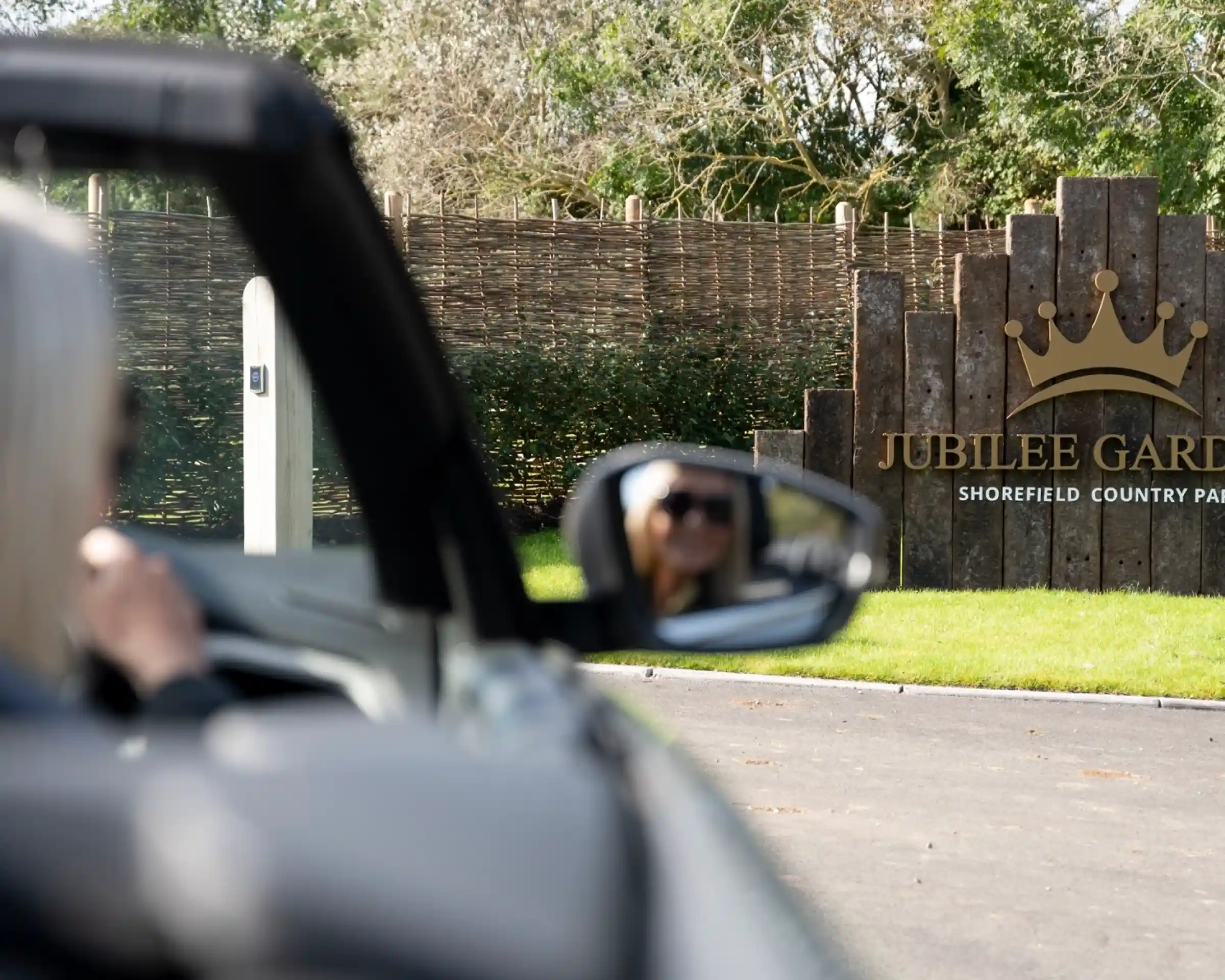 A person is seated in a convertible car, looking towards a sign that reads "Jubilee Gardens, Shorefield Country Park." The sign is surrounded by a wooden fence and green landscaping.