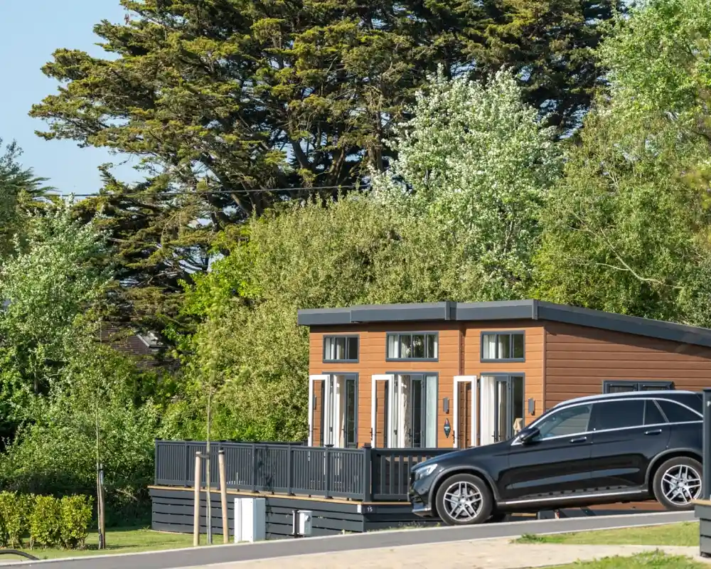 A modern wooden cabin with multiple windows and a spacious deck surrounded by lush greenery. A black SUV is parked nearby on a scenic road.