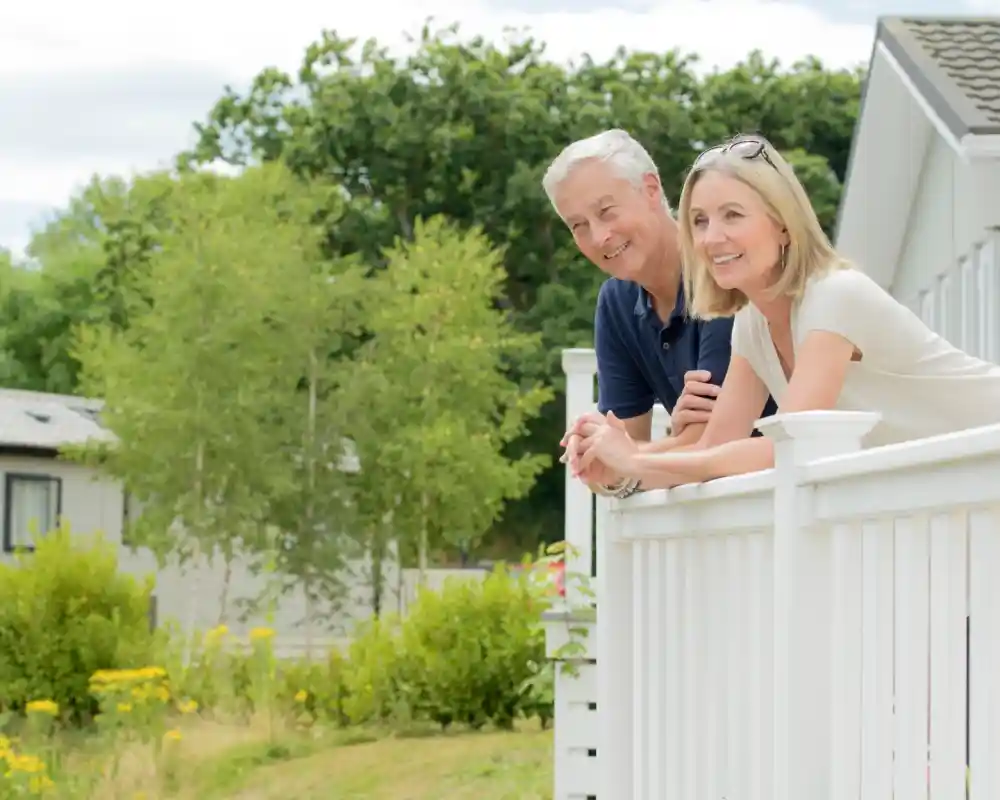 A smiling couple leaning on the railing of their holiday cabin, enjoying the view of the greenery around them.