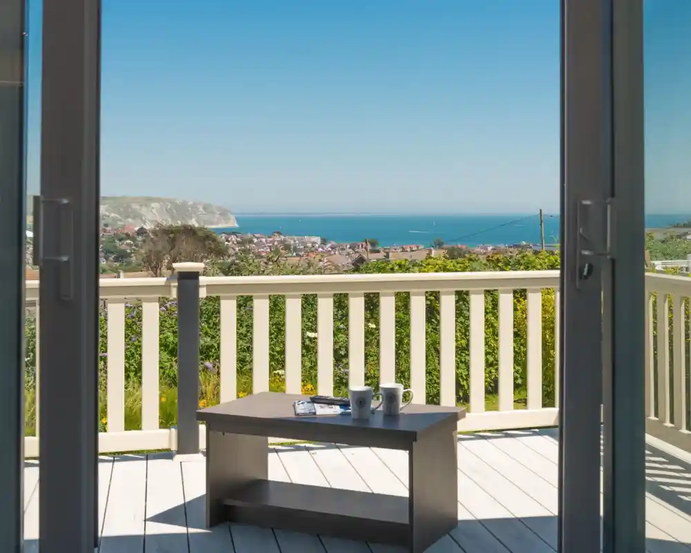 A view from inside a room, looking out through sliding glass doors onto a deck with a small table and two coffee cups. The bright blue ocean stretches into the distance, with a coastal town visible below. The scene is framed by the deck’s white railing, creating a peaceful, scenic atmosphere.