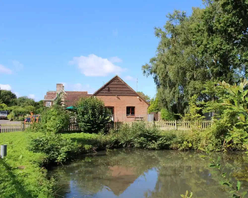 A charming brick house with a traditional gabled roof stands next to a small pond. Lush greenery surrounds the house, and there are trees both nearby and in the background. The house is enclosed by a wooden fence, and the sky is clear with a few clouds, giving the scene a peaceful, rural atmosphere.