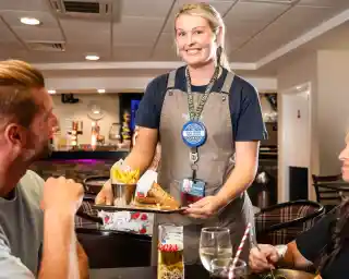 A waitress smiling while serving a plate of food with fries to two patrons at a restaurant table. A can of Coors beer is visible in front of one patron.