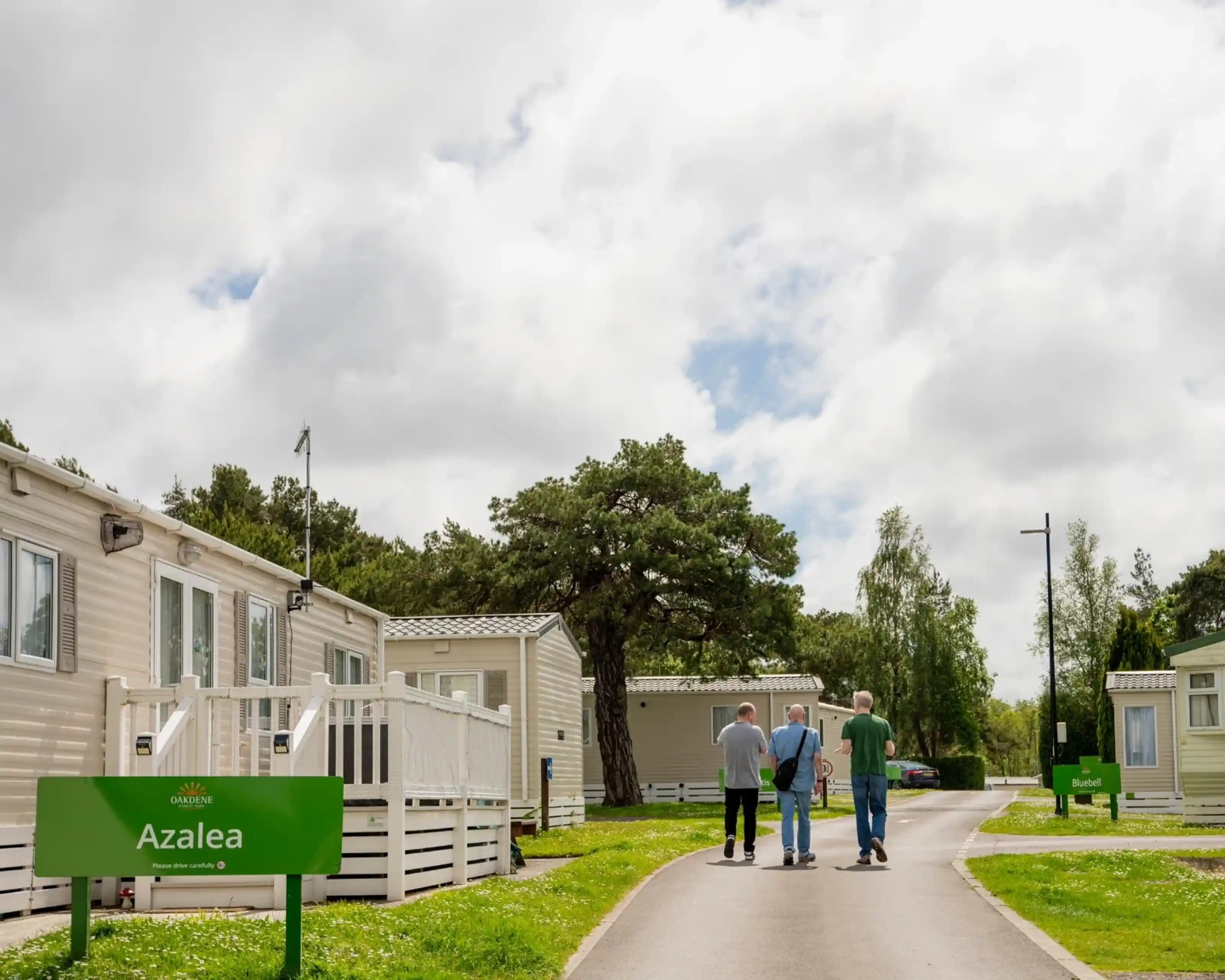Three people walking down a paved path in a mobile home park, flanked by white caravans and green trees. A sign that reads "Azalea" is visible on the left. The sky is partly cloudy.