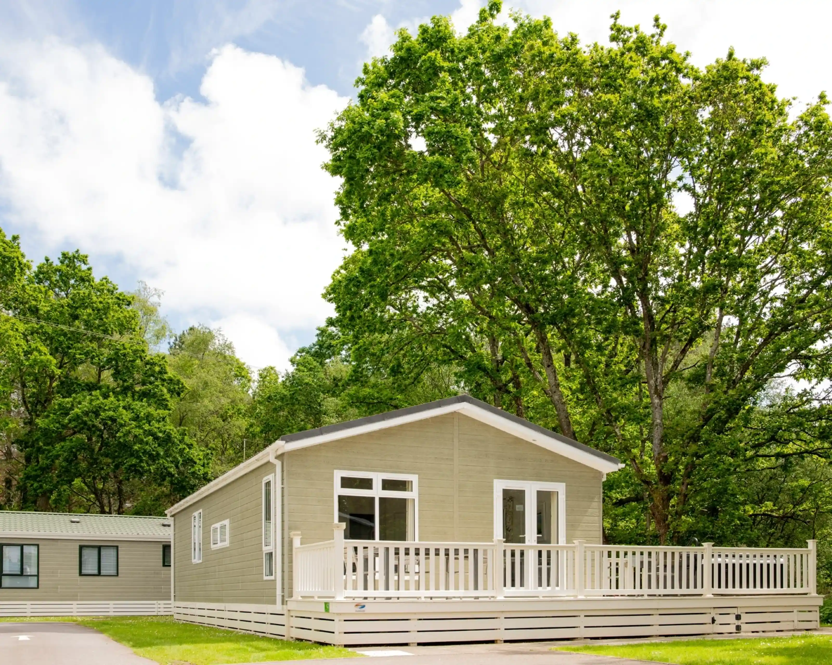 A modern wooden cabin with a front deck is surrounded by lush green trees and a blue sky with scattered clouds.