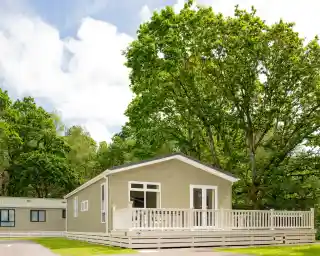 A modern wooden cabin with a front deck is surrounded by lush green trees and a blue sky with scattered clouds.