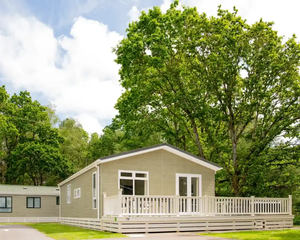 A modern mobile home with a light green exterior and a spacious wooden deck, surrounded by tall trees and greenery under a partly cloudy sky.