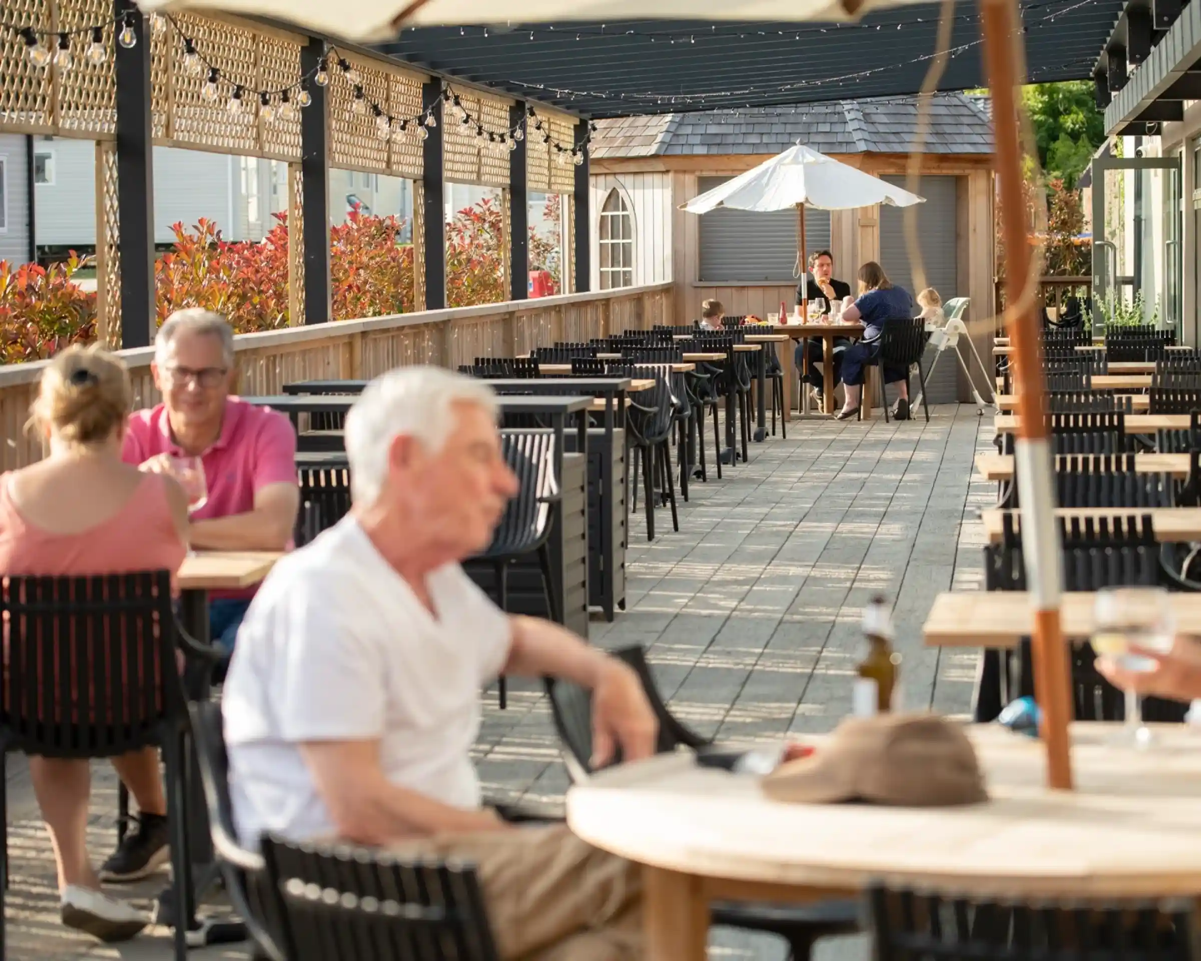 A outdoor dining area with wooden tables and chairs. Some patrons are seated while others are engaged in conversation. A white umbrella shades one table, and decorative lights are strung overhead. Lush greenery and colorful plants line the background.