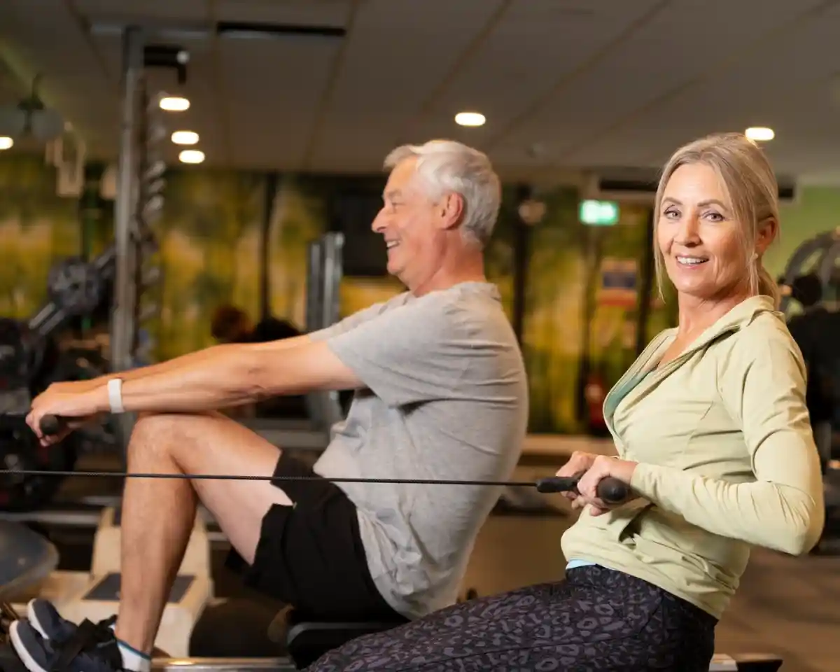 A man and a woman work out together at a gym, engaged in rowing exercises. The man is smiling and focused, while the woman looks confidently at the camera. They are dressed in comfortable athletic clothing, with weights and gym equipment in the background.