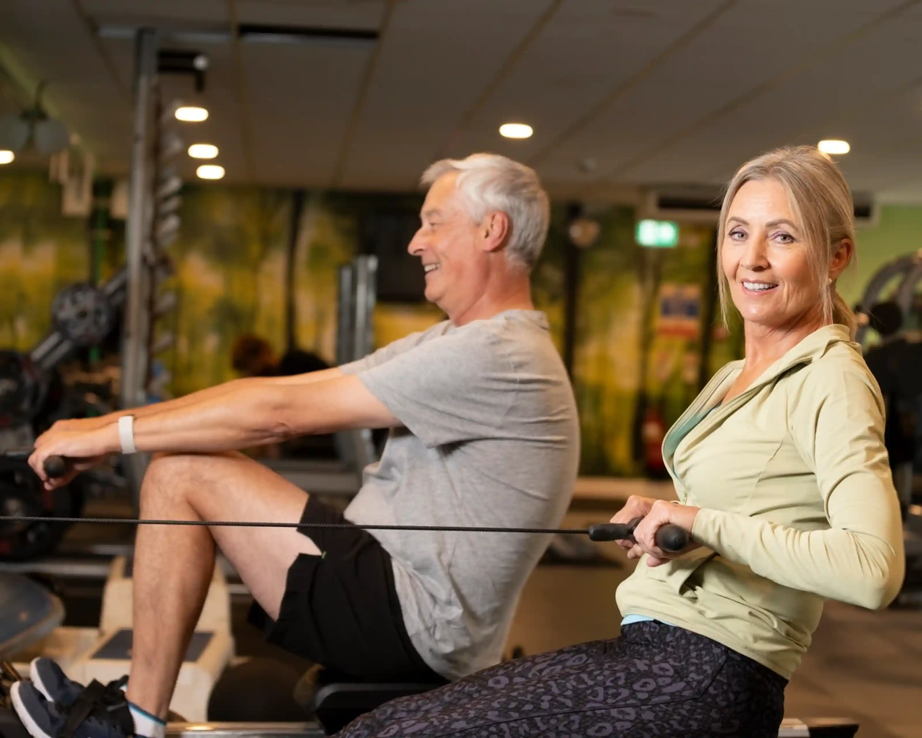 A man and a woman work out together at a gym, engaged in rowing exercises. The man is smiling and focused, while the woman looks confidently at the camera. They are dressed in comfortable athletic clothing, with weights and gym equipment in the background.