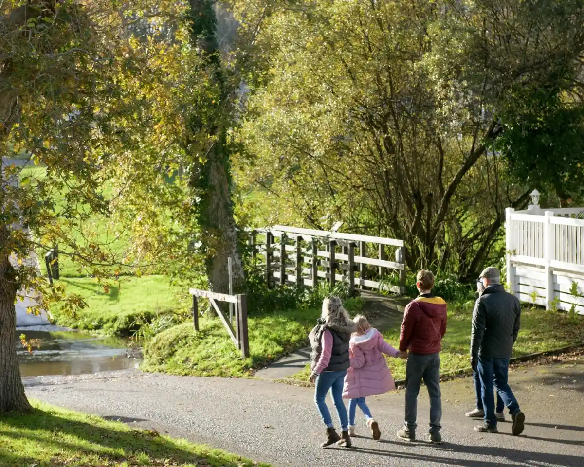 A family walks along a path in a sunny park, surrounded by trees and greenery. The adults are wearing jackets, and a child is in a pink coat, holding hands with one of them. A wooden bridge is visible in the background.
