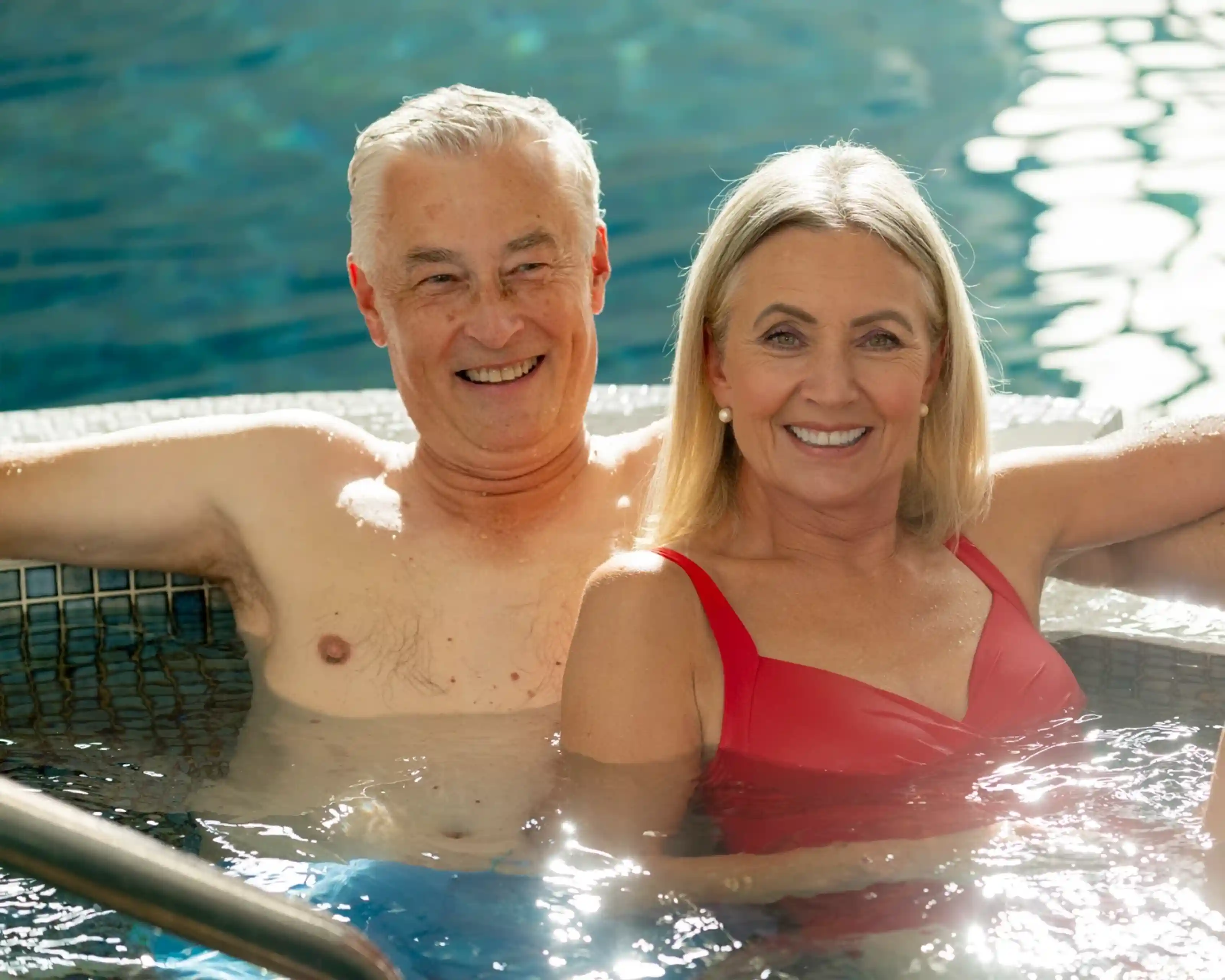 A smiling couple relaxes in a hot tub, surrounded by water and soft natural light. The man has gray hair and is shirtless, while the woman has blonde hair and is wearing a red swimsuit. They appear happy and comfortable together.