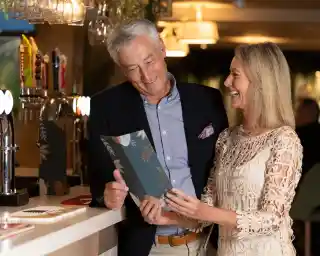 A smiling older couple stands together at a bar, looking at a menu. The man, dressed in a dark blazer and light shirt, has gray hair and is laughing. The woman, in an elegant lace dress, is also smiling as she engages with him. The background features bar taps and warm lighting.