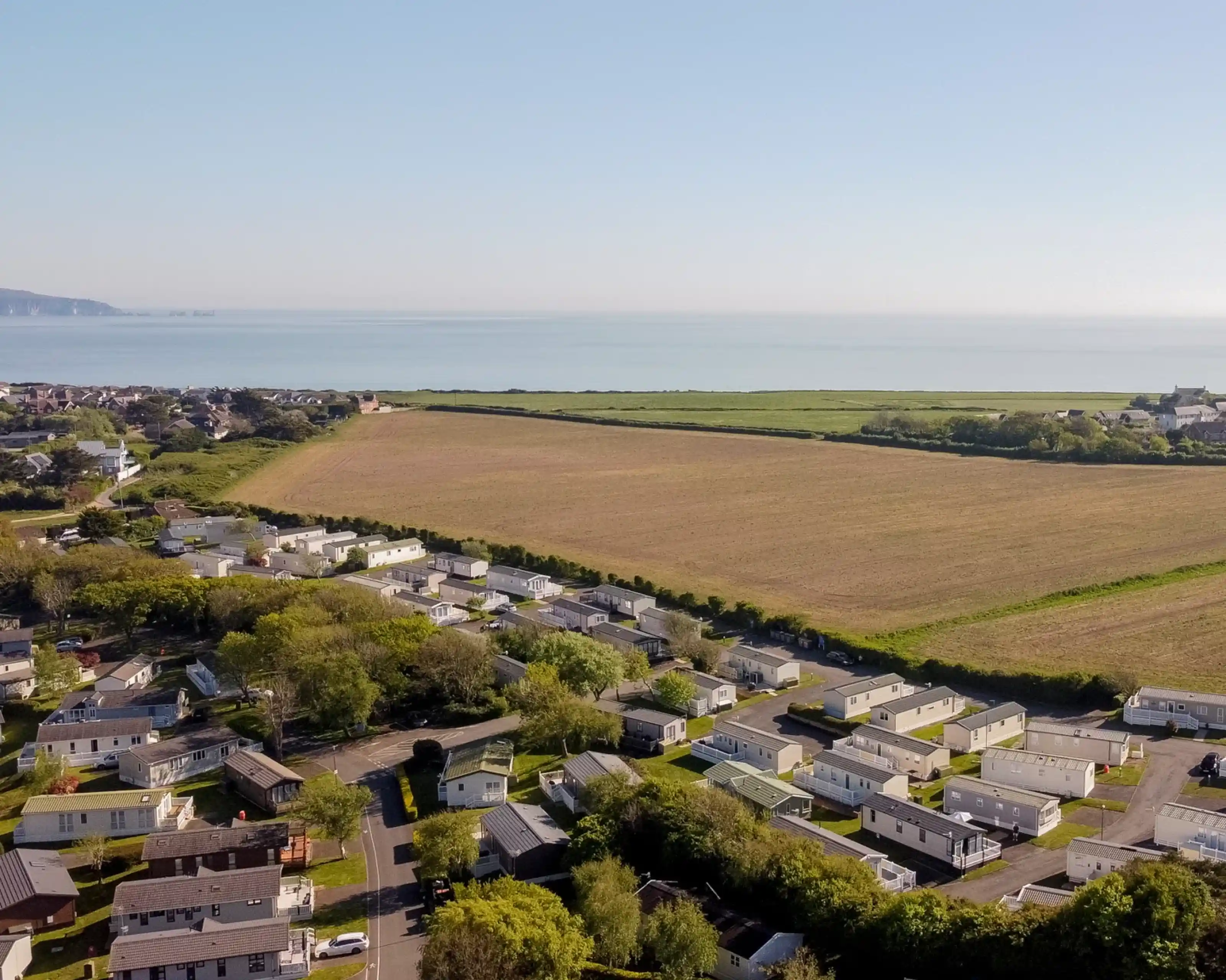Aerial view of a coastal area featuring a residential park with numerous trailers, a wide field, and the ocean in the background under a clear blue sky.