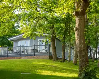 A modern gray cabin nestled among lush green trees, featuring a spacious deck that overlooks a gently sloping lawn. Sunlight filters through the leaves, creating a warm and inviting atmosphere.